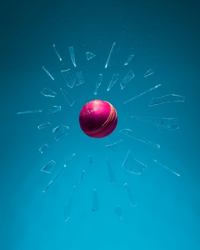 Dynamic shot of a red cricket ball breaking through glass against a teal backdrop.